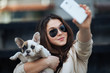 Beautiful young girl smiling and taking a selfie with her cute French bulldog puppy. Urban scene. Selective focus on girl.