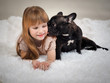 The dog's licking the baby. Redhead little girl and a pet dog - black French bulldog